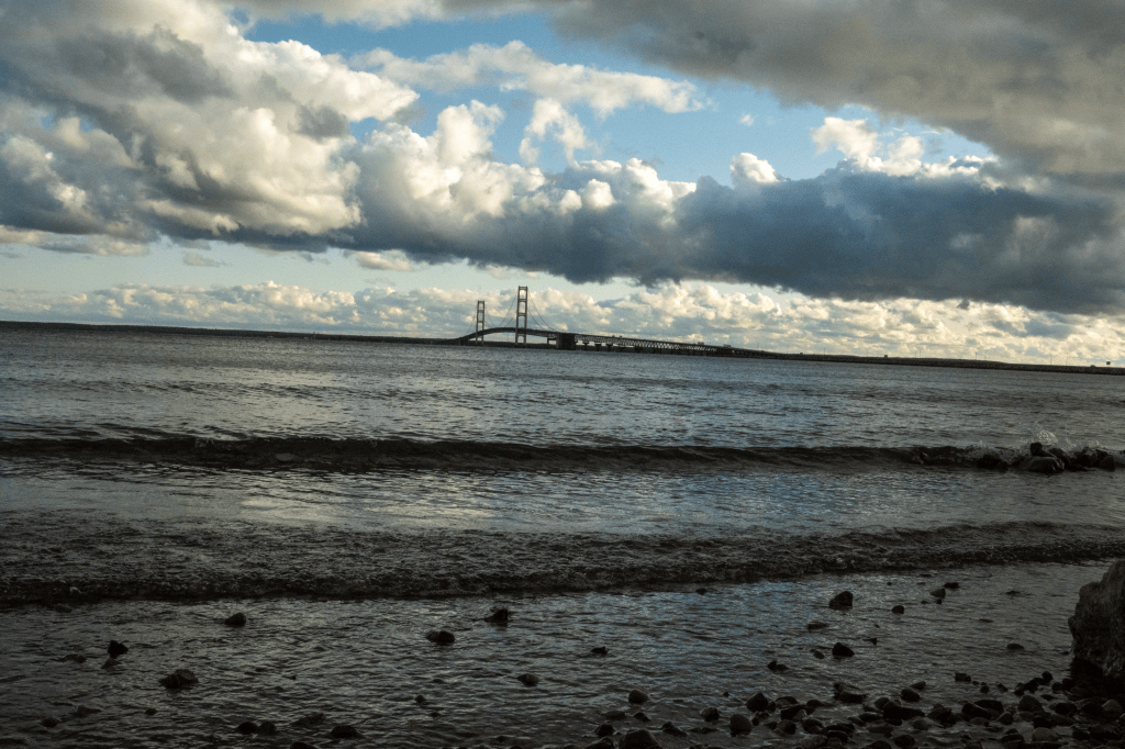 Clouds with a blue sky showing over light waves rolling against the shore in front of the Mackinac Bridge