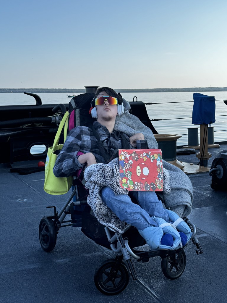 Tristan sitting in his wheelchair on the deck of a coast guard ship with his iPad out to pass a little bit of time.
